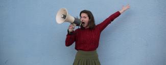 jeune femme portant un pull rouge et criant dans un mégaphone devant un mur bleu-gris