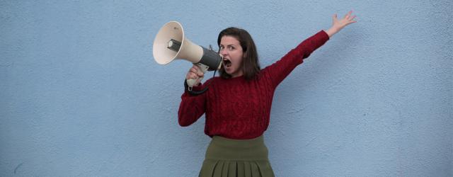 Femme portant un pull rouge, hurlant dans un mégaphone devant un mur bleu